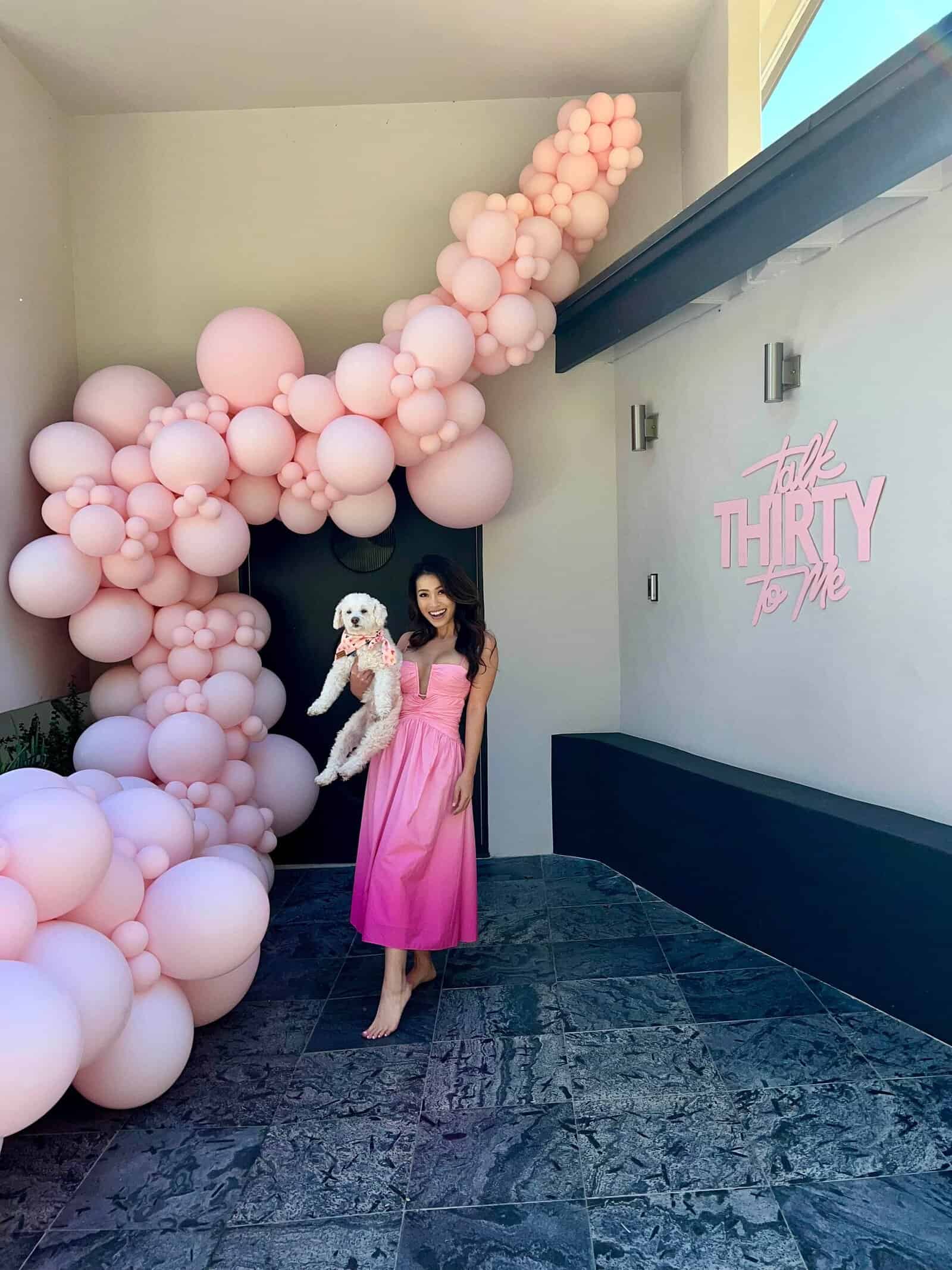 Woman in a pink dress holds a small white dog, standing by an elegant pink balloon installation and a wall sign that reads "talk THIRTY to me"—a picture-perfect moment in Los Angeles.