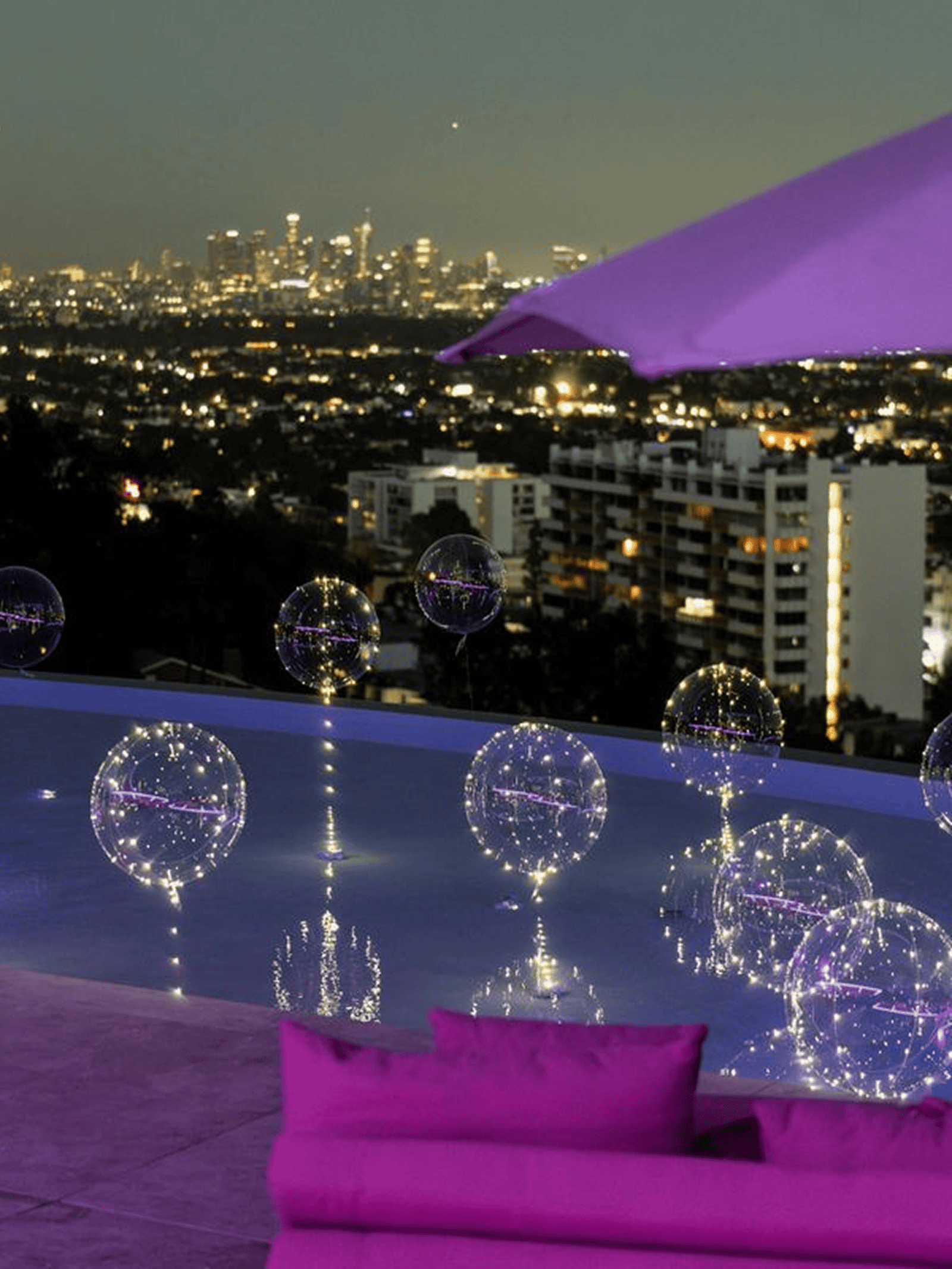 A rooftop pool at dusk in Los Angeles with glowing orbs and balloons floating on the water, purple seating and umbrella, set against a stunning city skyline—perfect for a memorable corporate event.