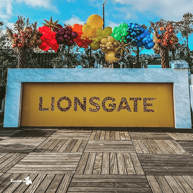 A marble counter with a yellow panel displays the word "LIONSGATE" in floral letters, backed by vibrant balloon decor from a skilled balloon artist and lush greenery outdoors.