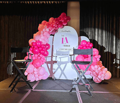 Two director chairs sit on a stage in Los Angeles, adorned with stunning balloon decor and a sign reading "The LA Woman – A Power Panel Event.