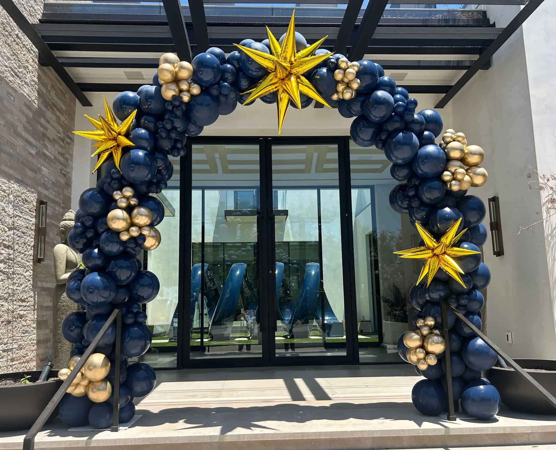 A decorative balloon installation of navy blue and gold balloons, featuring three gold star-shaped balloons, stands in front of glass double doors—perfect for a stylish corporate event.