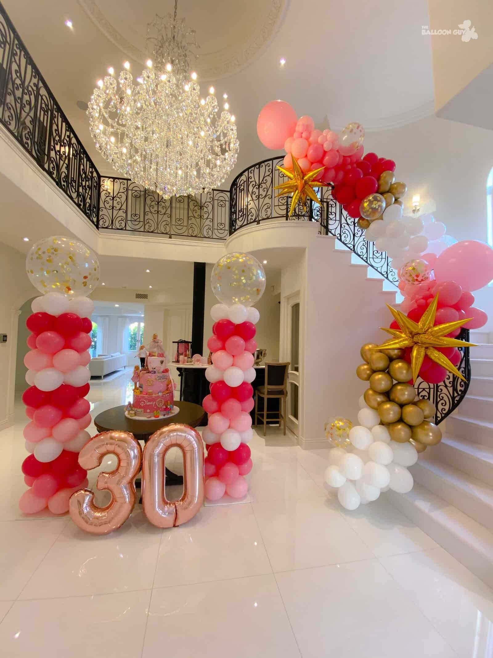 A decorated foyer in Los Angeles features pink, white, and gold balloons, a striking "30" balloon display, a cake on the table, and a chandelier overhead—perfect for an elegant 30th birthday celebration.