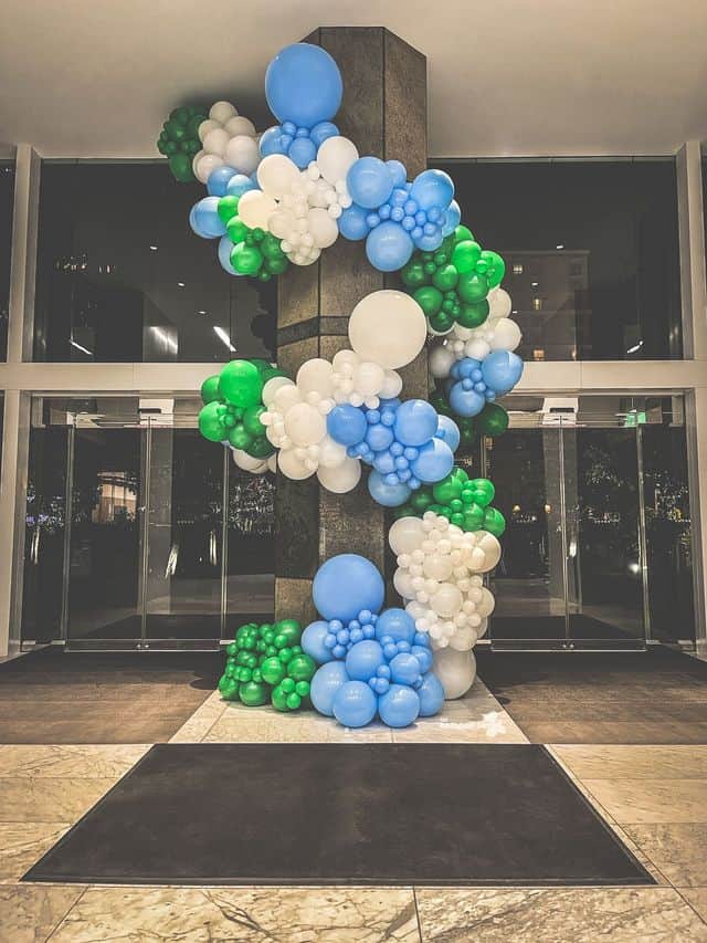 Clusters of blue, white, and green balloon decor are arranged around a pillar in a modern building lobby with glass doors in the background.