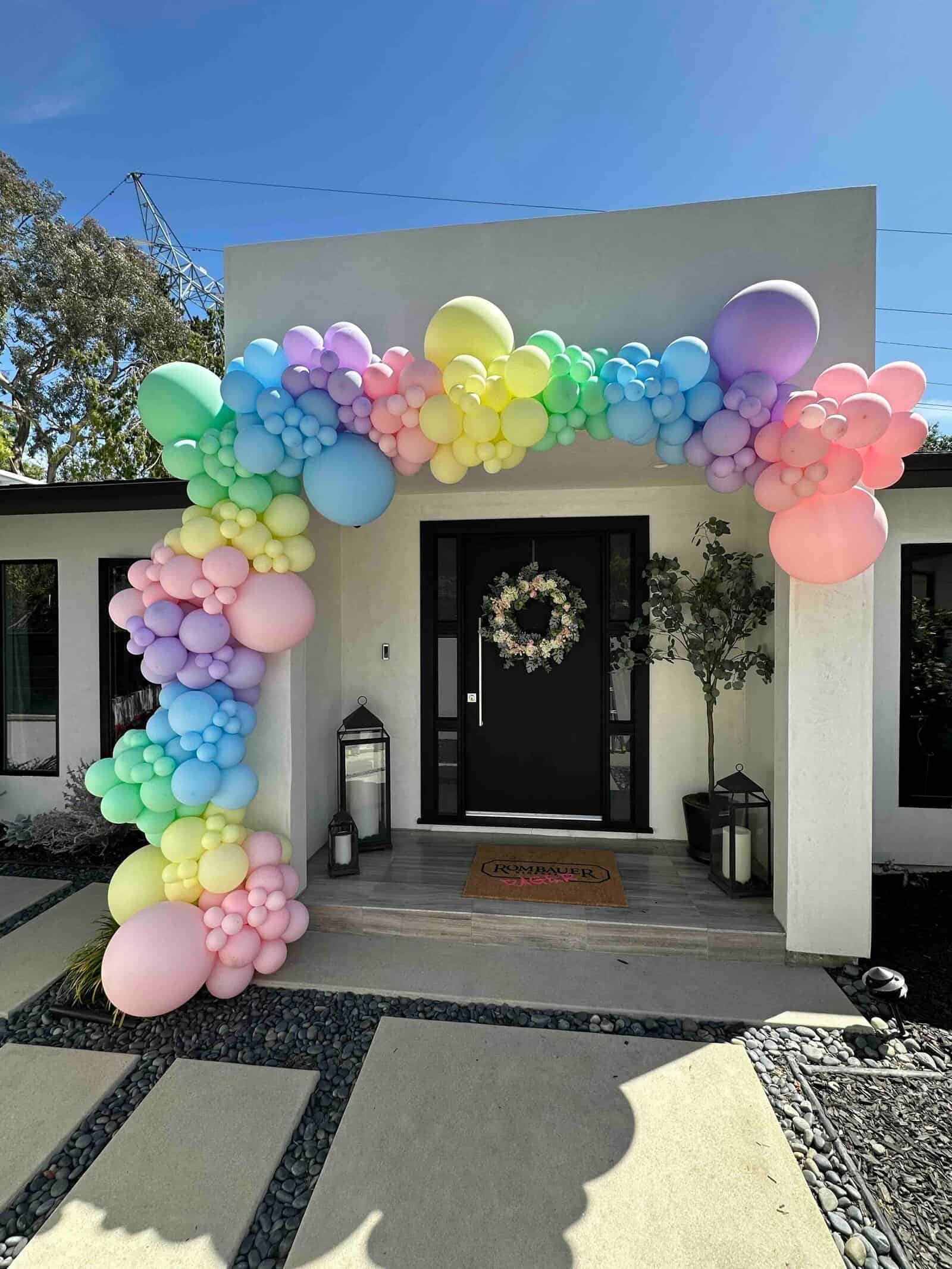 A pastel rainbow balloon arch by a talented balloon artist is set up over the entrance of a modern Los Angeles house with a black door and wreath, flanked by large lanterns.