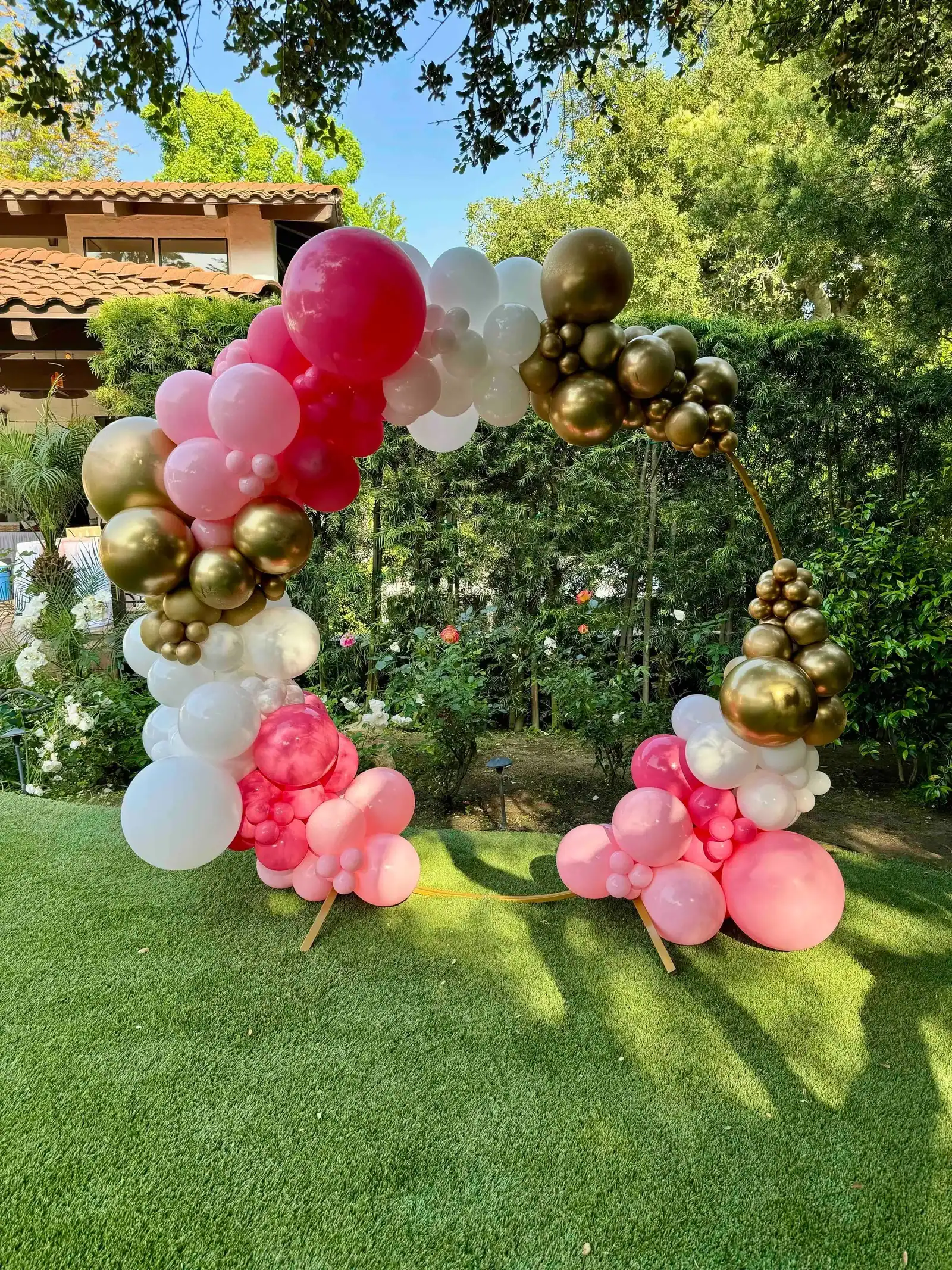 A circular arch featuring stunning balloon decor in pink, white, and gold stands on a green lawn with trees and a house in the background.