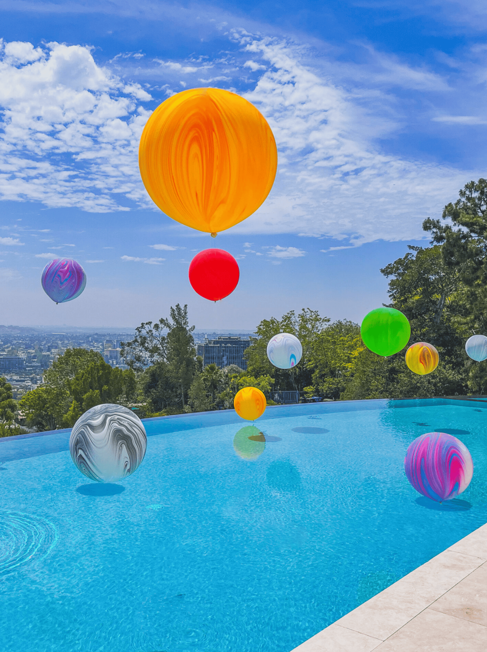 Large colorful balloon installations float above a clear blue infinity pool on a sunny day, with trees and cityscape visible in the background—perfect for stylish corporate event balloon decor.