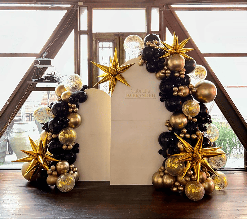 Two balloon pillars in black, gold, and clear with gold star accents flank a white backdrop inside a bright room with large windows—elegant balloon decor perfect for any corporate event.