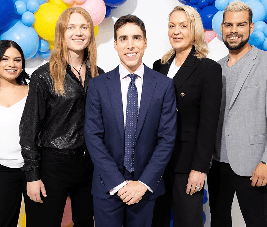 Five people pose together in front of vibrant balloon decor. Four are dressed in business or business-casual attire, while one stands out in a shiny black shirt.