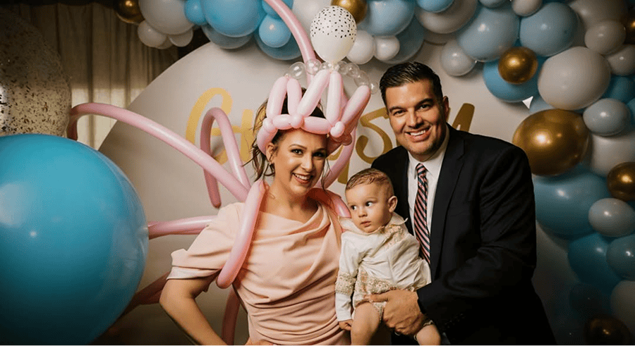 Three people pose in front of a festive balloon arch installation; the woman wears a pink balloon hat, the man is in a suit, and they are holding a young child dressed in light-colored clothes.