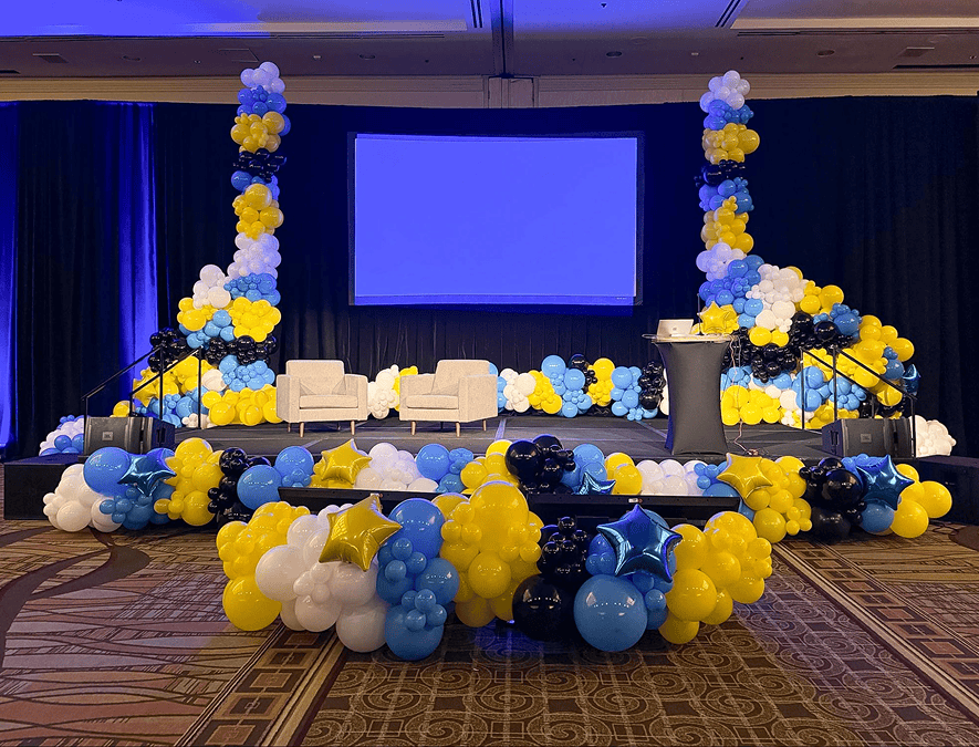 A stage decorated with vibrant balloon decor in clusters of blue, yellow, white, and black balloons, two white chairs, a podium, and a large blank screen in the background.