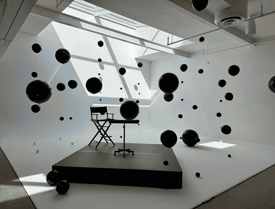 A director's chair and table sit on a platform in a white Los Angeles studio, surrounded by suspended black spheres, reminiscent of décor for a corporate event, all under a large skylight.