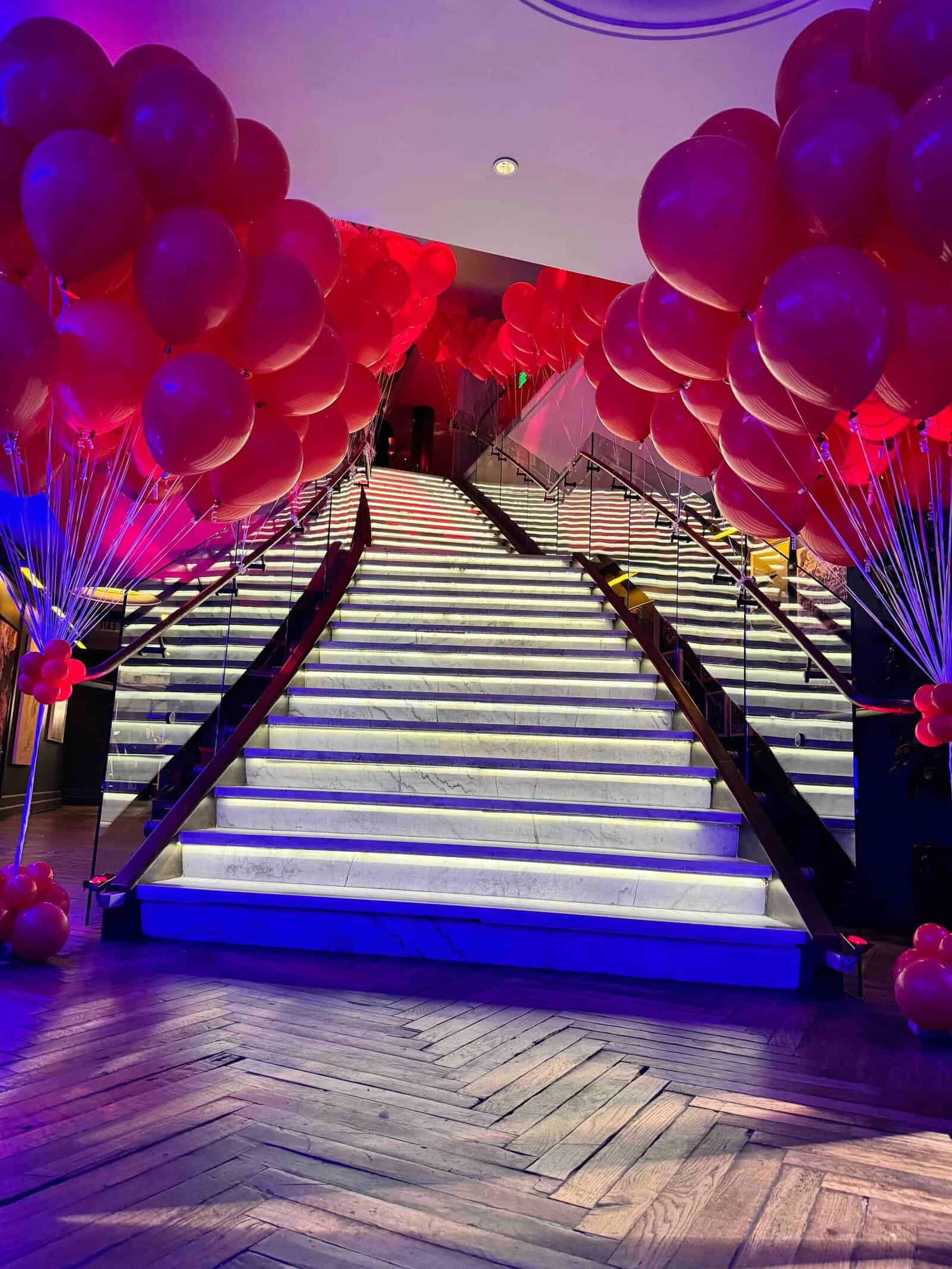 A wide marble staircase with illuminated steps, flanked by striking balloon decor from a skilled balloon artist, sets the scene for an elegant corporate event in a beautifully decorated indoor space.
