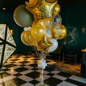 The Signature Balloon Bouquet, featuring gold, white, green, and transparent balloons—plus gold star-shaped ones—creates a striking installation on a checkered black and white floor in a modern room.