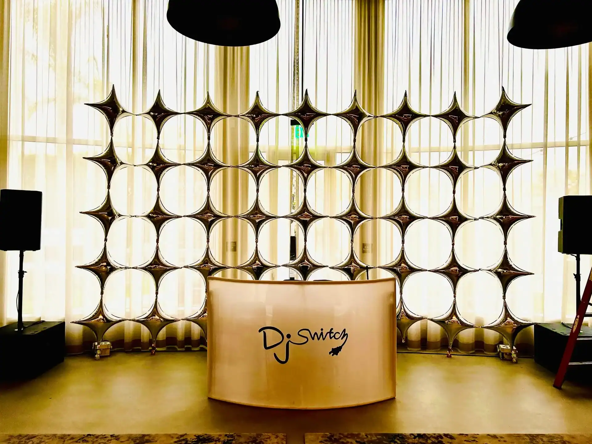 A DJ booth labeled "DJ Switch" is set up in front of striking balloon installations with a metallic backdrop, large speakers on either side, and sheer curtains in the background.