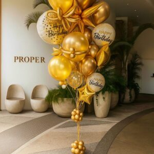 The Luxe gold and white "Happy Birthday" balloon cluster, featuring star shapes, brightens a modern Los Angeles lobby with curved seating and potted plants.