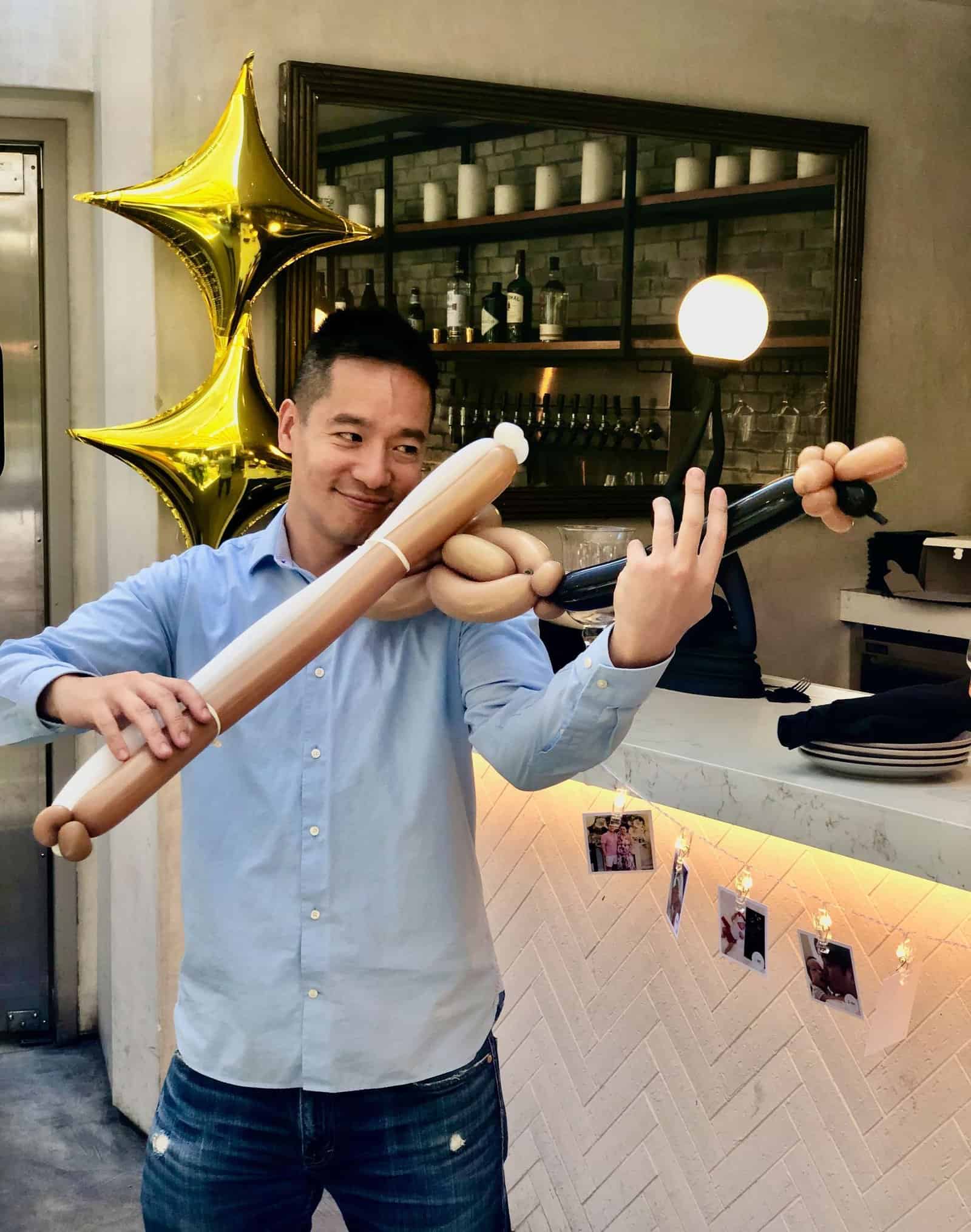 A man in a blue shirt pretends to play a violin made of balloons, standing near a counter with creative balloon decor and star-shaped balloons, with hanging photos in the background at an event in Los Angeles.