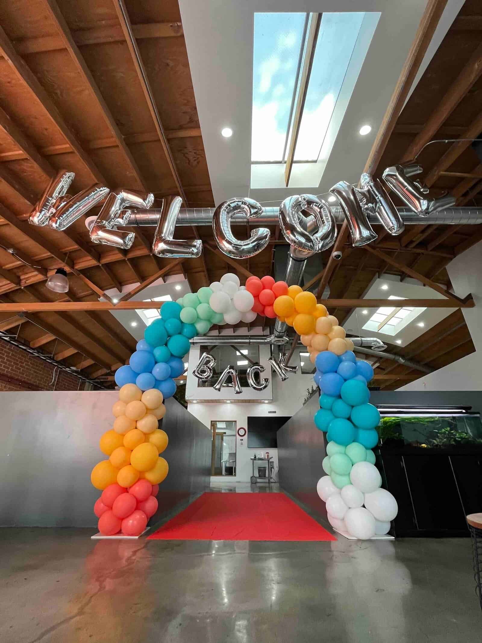 A balloon arch and metallic "WELCOME BACK" balloons, crafted by a skilled balloon artist, are displayed above a red carpet in an office space with exposed wooden beams and skylights.