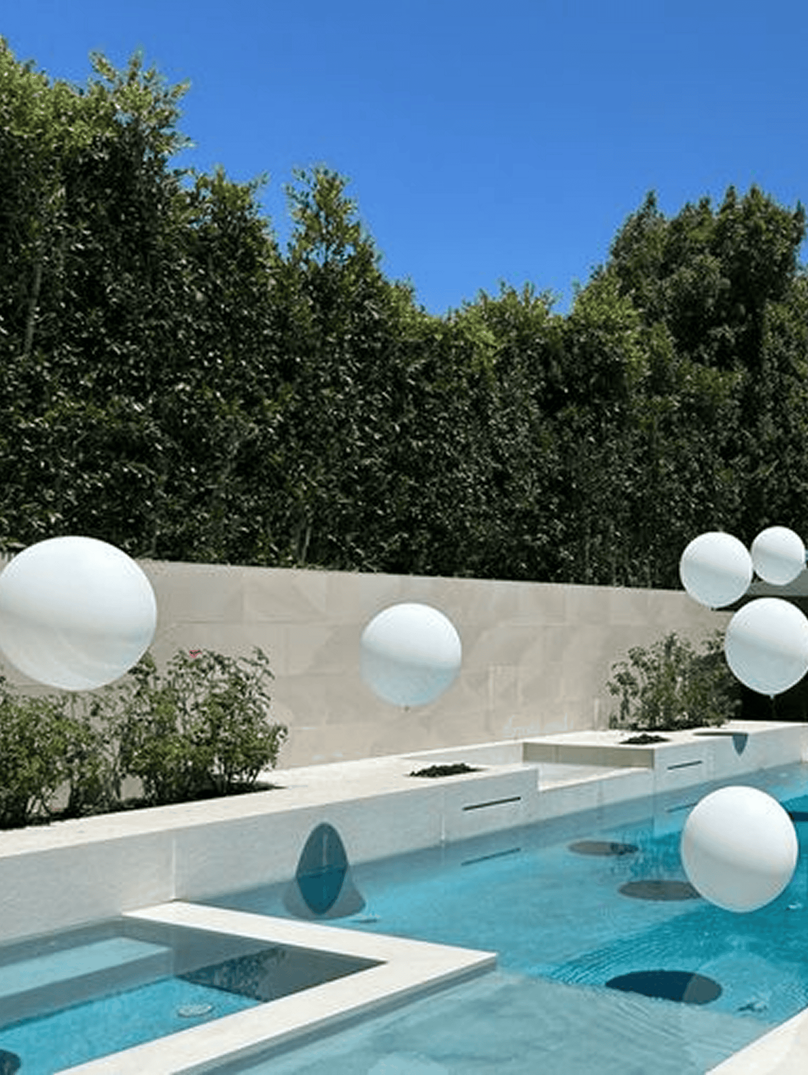 Modern swimming pool with clear water, surrounded by white tiles and tall green hedges, features several large white balloons arranged by a balloon artist, floating elegantly above the water.