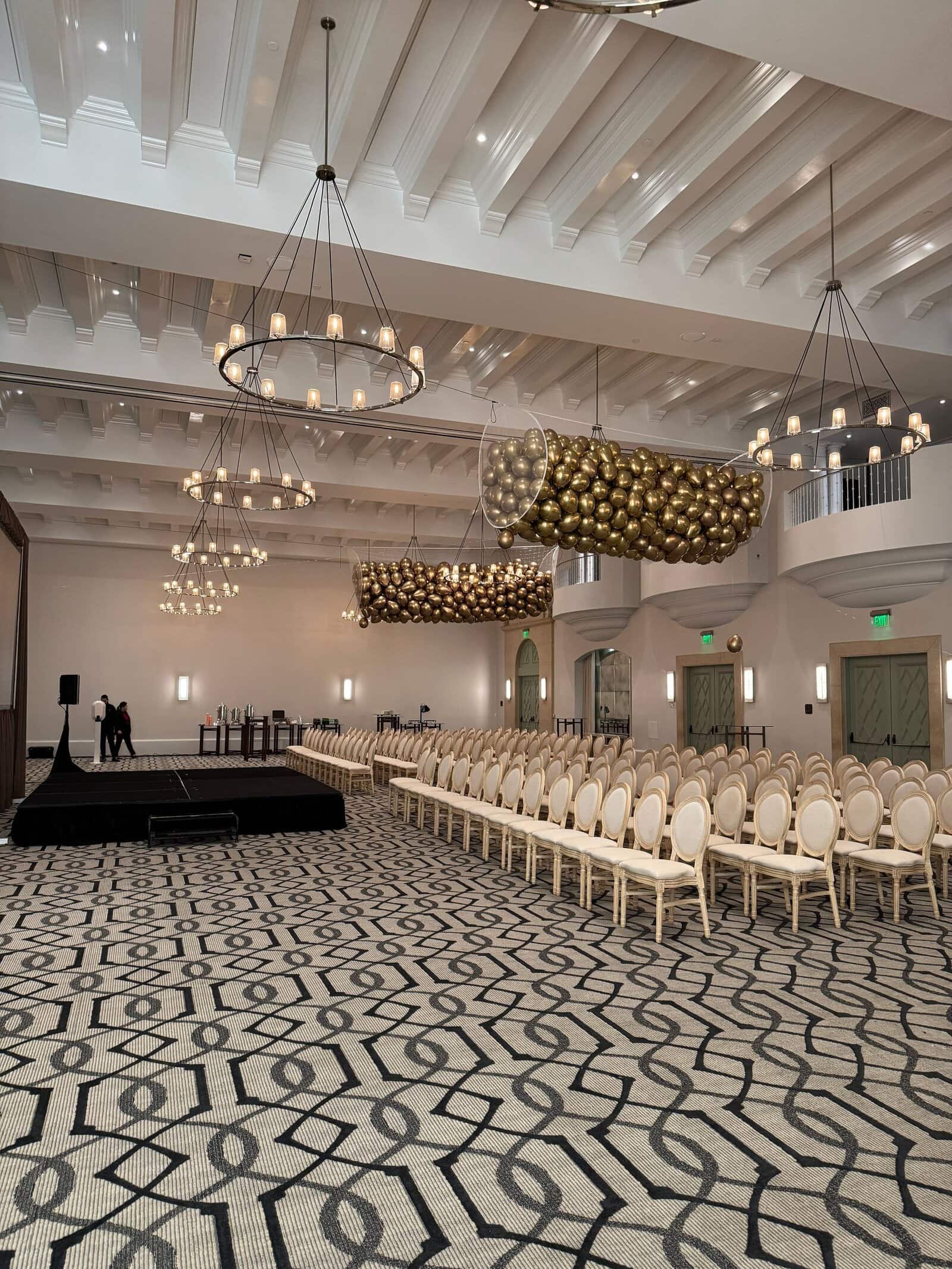 A large, empty event hall with rows of cream-colored chairs, a patterned carpet, chandeliers, and a stage set up at one end—perfect for a corporate event or hosting a talented balloon artist.