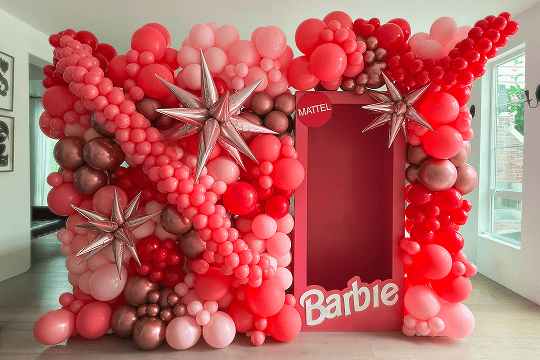 A life-sized Barbie doll box surrounded by vibrant balloon installations featuring pink, red, and metallic balloons with silver star-shaped decorations, creating stunning indoor balloon decor.