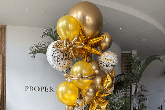 A bouquet of gold and white balloon decor, some with "Happy Birthday" messages and star shapes, arranged indoors near a wall with the word "PROPER" visible—perfect for stylish balloon installations in Los Angeles.