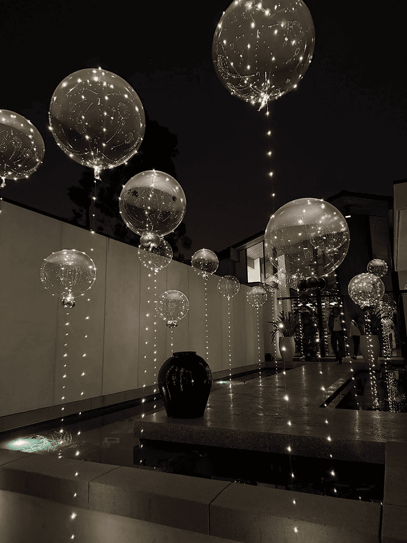 A modern outdoor space at night features large transparent balloons filled with string lights, floating above a reflecting pool with minimalist decor—perfect for a stunning corporate event or elegant balloon installations.