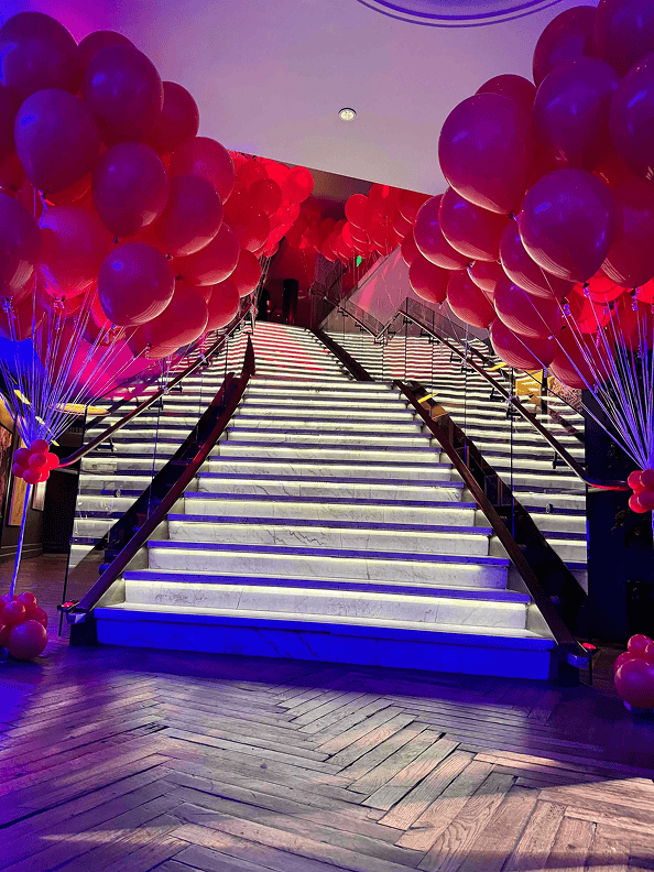 A grand staircase with illuminated steps is adorned with stunning balloon decor by a skilled balloon artist, featuring vibrant red balloons on both sides and creating a festive atmosphere in a modern indoor setting.