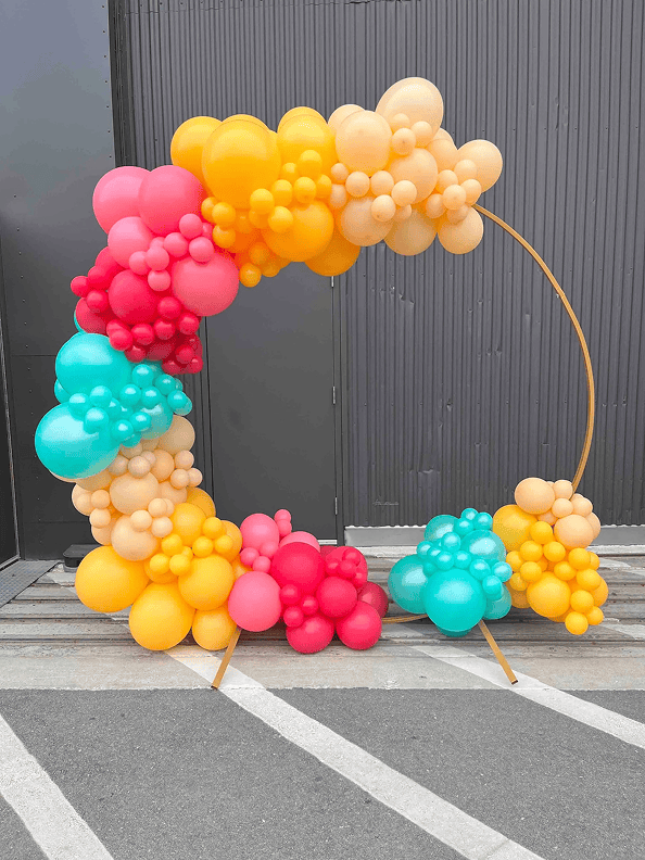 A semicircular metal frame decorated with clusters of red, yellow, orange, and teal balloon decor stands outdoors on a concrete surface.