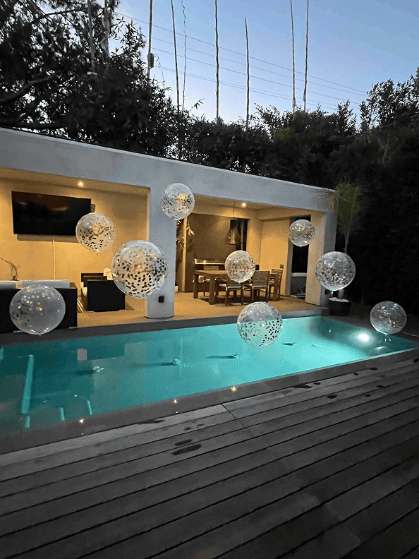 Modern backyard with a rectangular pool, wooden deck, and a covered patio area; decorative balloons from a skilled Los Angeles balloon artist float above the pool at dusk.
