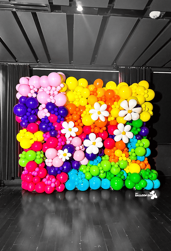 A colorful balloon wall display featuring rows of balloons in various colors and sizes, decorated with large white balloon flowers by a talented balloon artist—perfect for any corporate event.