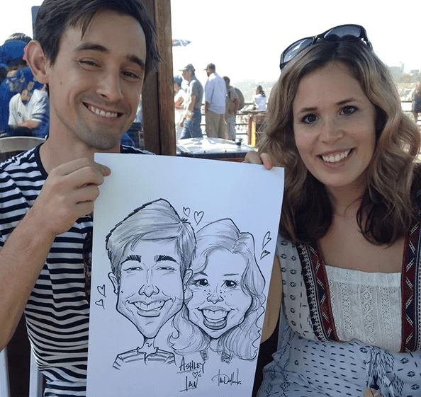 A man and a woman sit side by side outdoors, smiling and holding up a black-and-white caricature drawing of themselves, with vibrant balloon decor in the background.