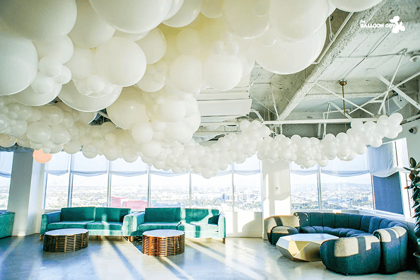 A modern lounge with teal sofas and round tables in Los Angeles, featuring large clusters of white balloons suspended from the ceiling.