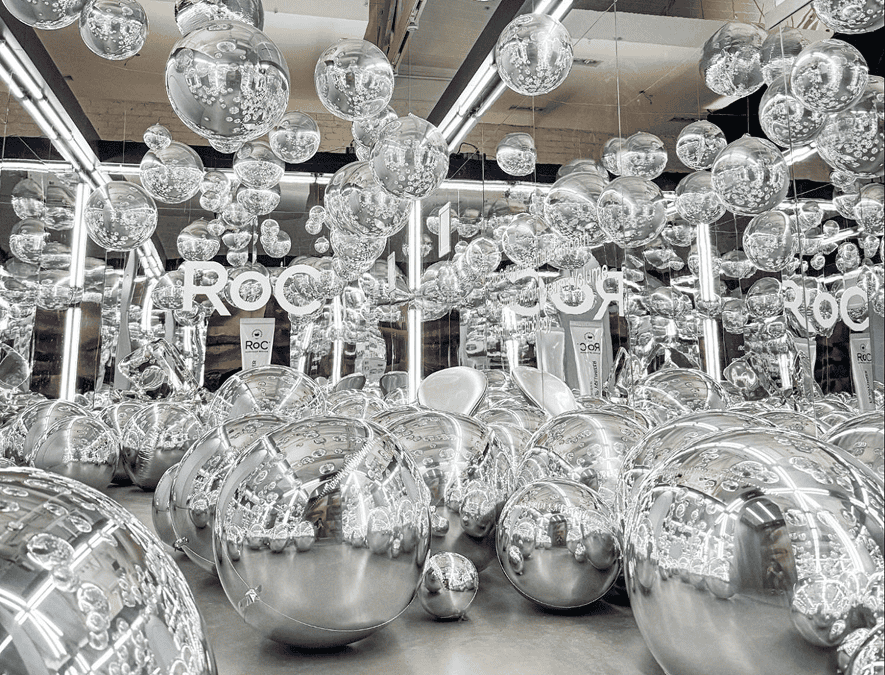 Room filled with various sizes of reflective silver spheres, like a stunning balloon installation, suspended and on the ground, with bright vertical lights and "ROC" signs in the background—perfect for a stylish Los Angeles corporate event.