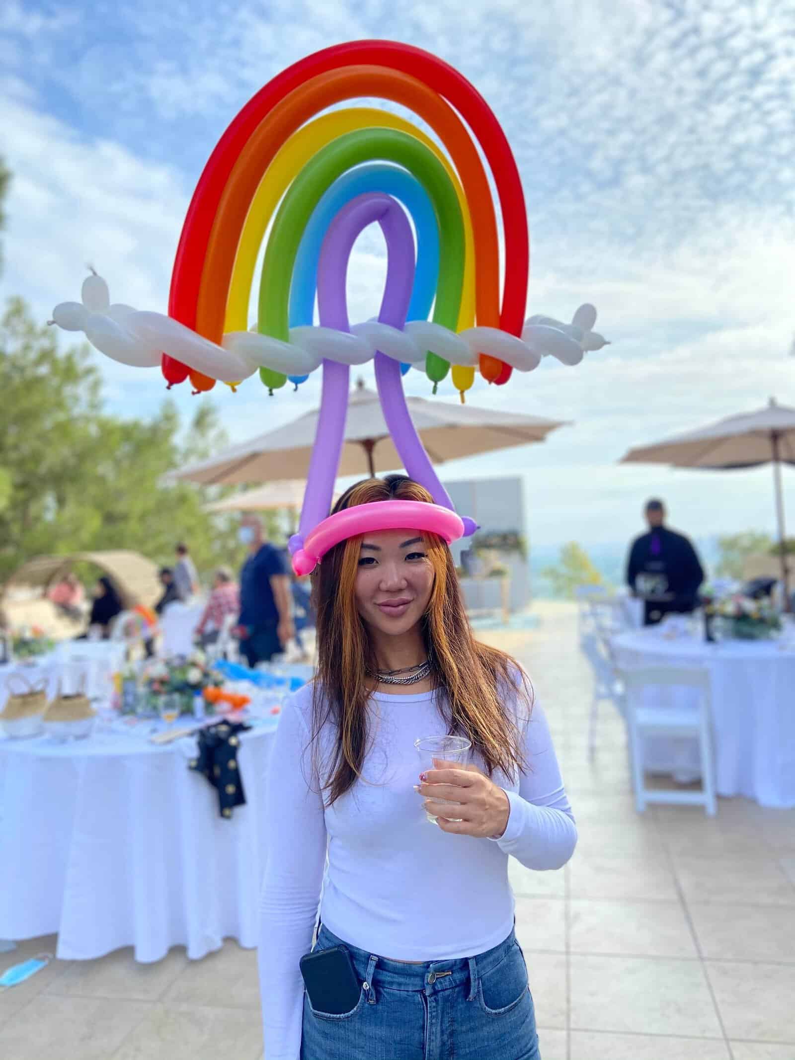 A woman wearing a large rainbow balloon hat stands outdoors at a Los Angeles event, holding a drink, with festive balloon decor, tables, and people in the background.