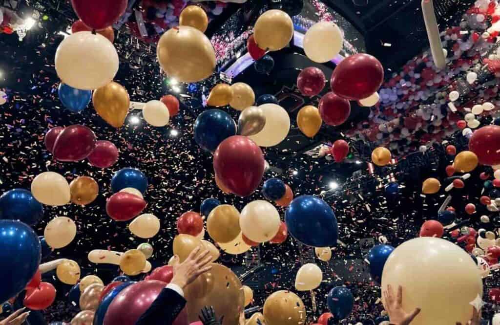 Colorful balloon drops and confetti fall from the ceiling as people raise their hands, creating a festive atmosphere with lively event decor during an indoor celebration.