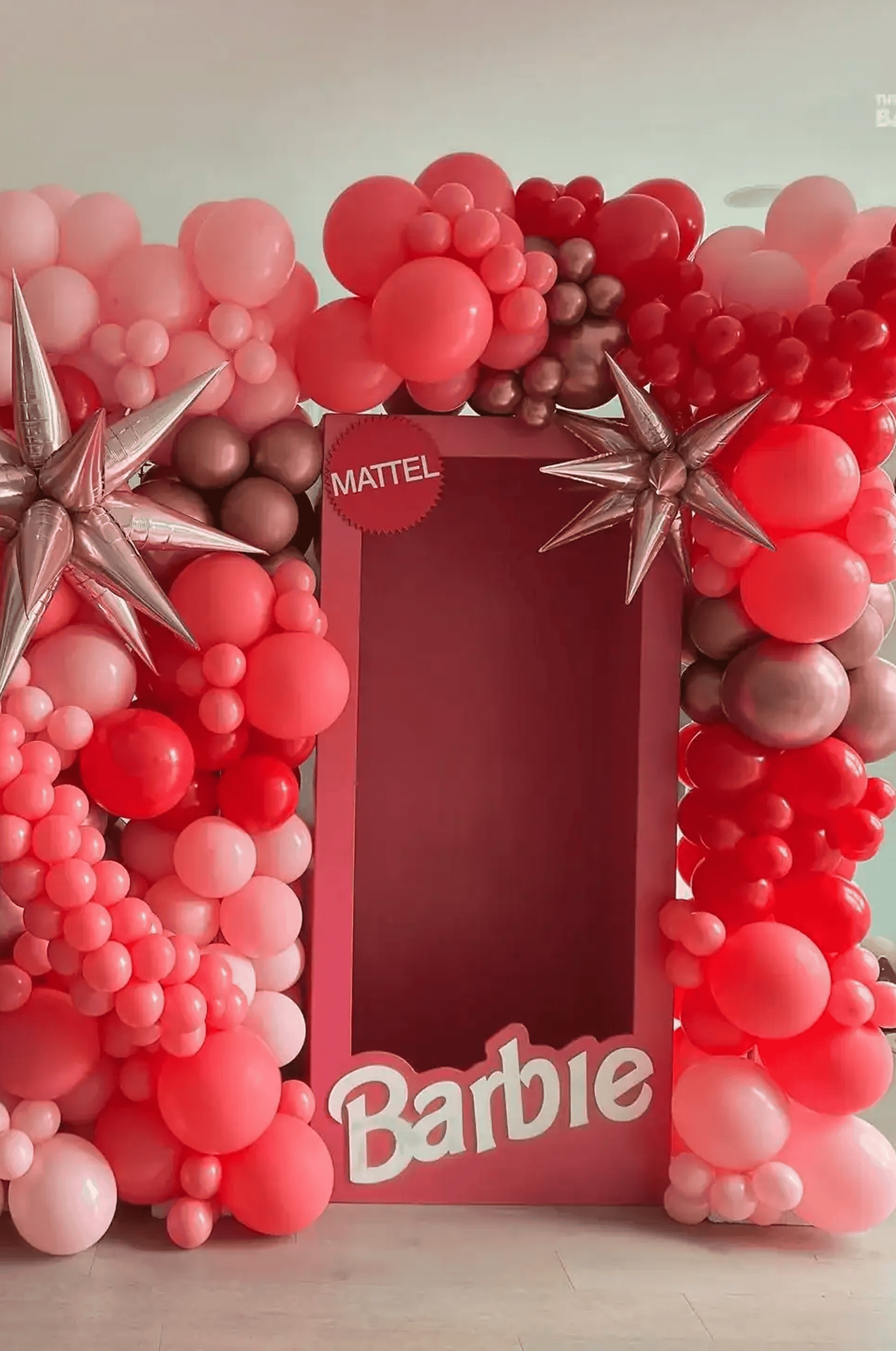 A large pink and red balloon arch, perfect for event decor, surrounds a cardboard Barbie doll box frame with the Mattel logo, designed as a photo booth.