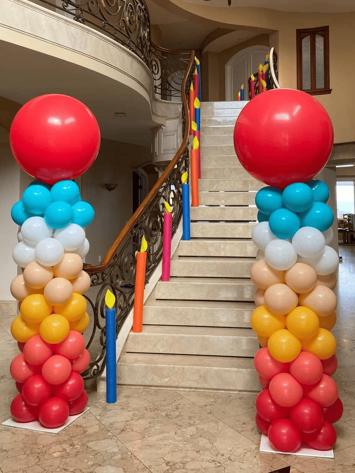 Two tall balloon columns made of colorful balloons and topped with large red balloons stand at the base of a curved staircase adorned with balloon “candles,” creating eye-catching event decor.