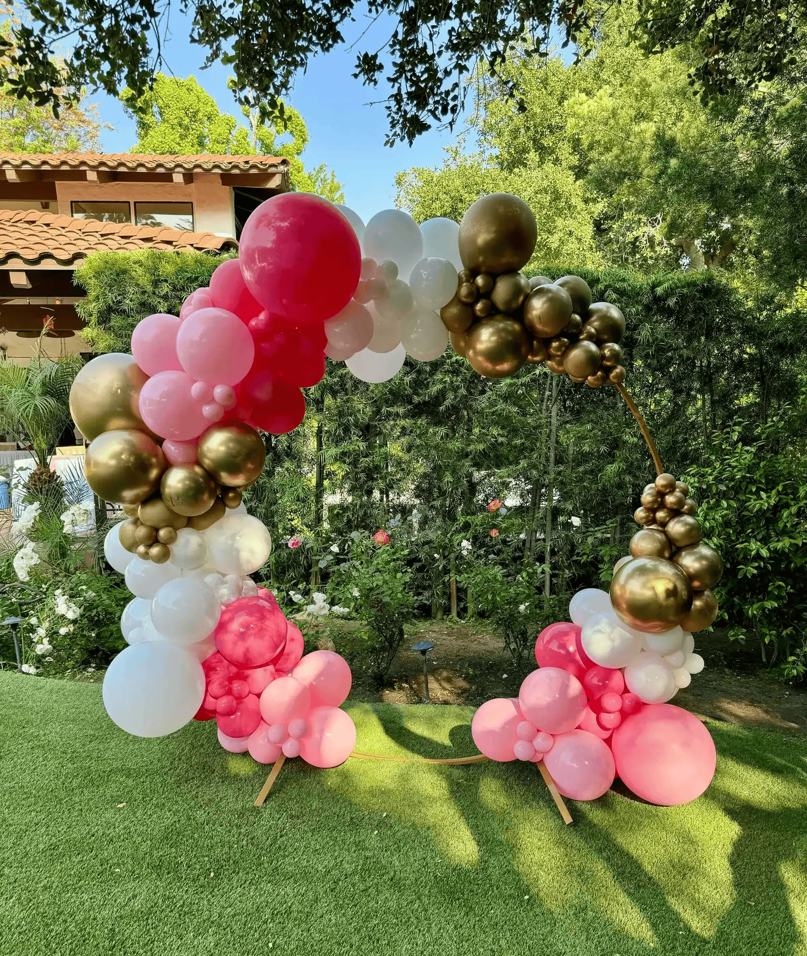 A circular balloon arch on a sturdy balloon stand features pink, white, and gold balloons, standing elegantly on grass in a garden with trees and a house as the backdrop—perfect for a charming party decoration.