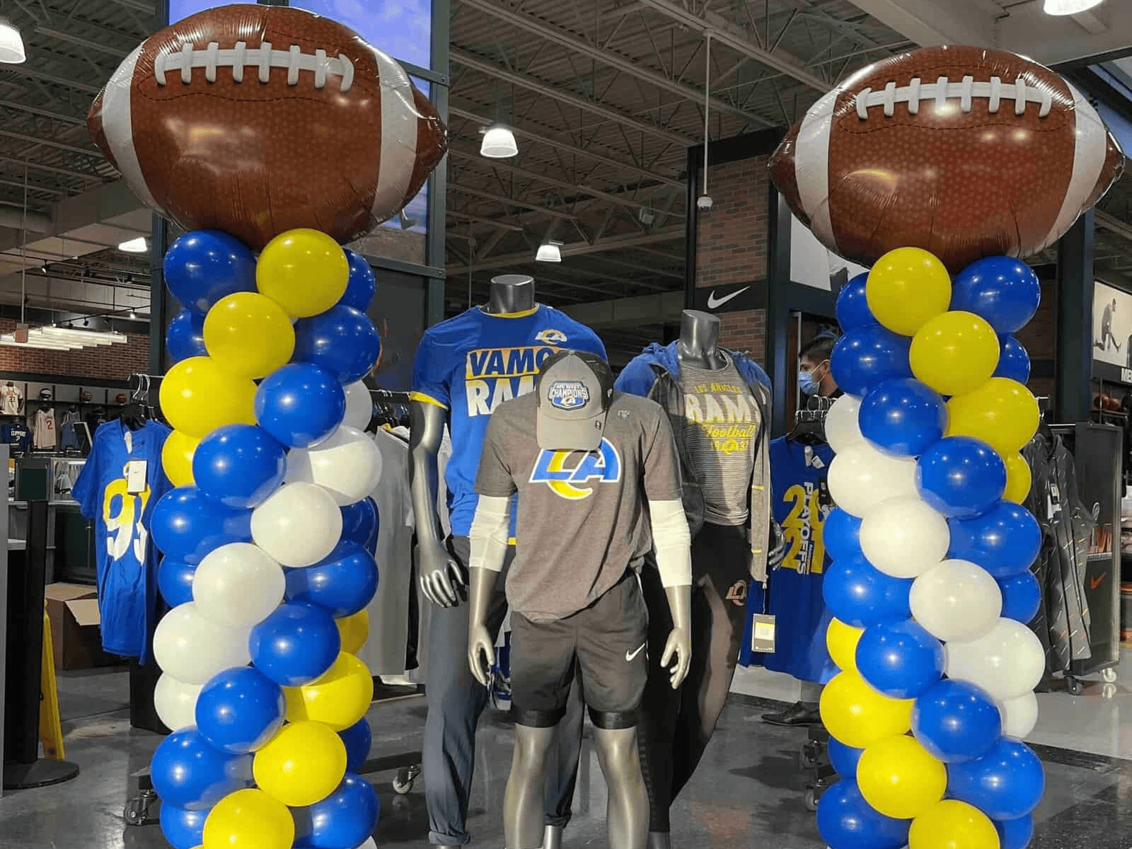 Store display with mannequins dressed in Los Angeles Rams gear, perfect for Sports Events, flanked by blue, yellow, and white balloon columns topped with large inflatable footballs.