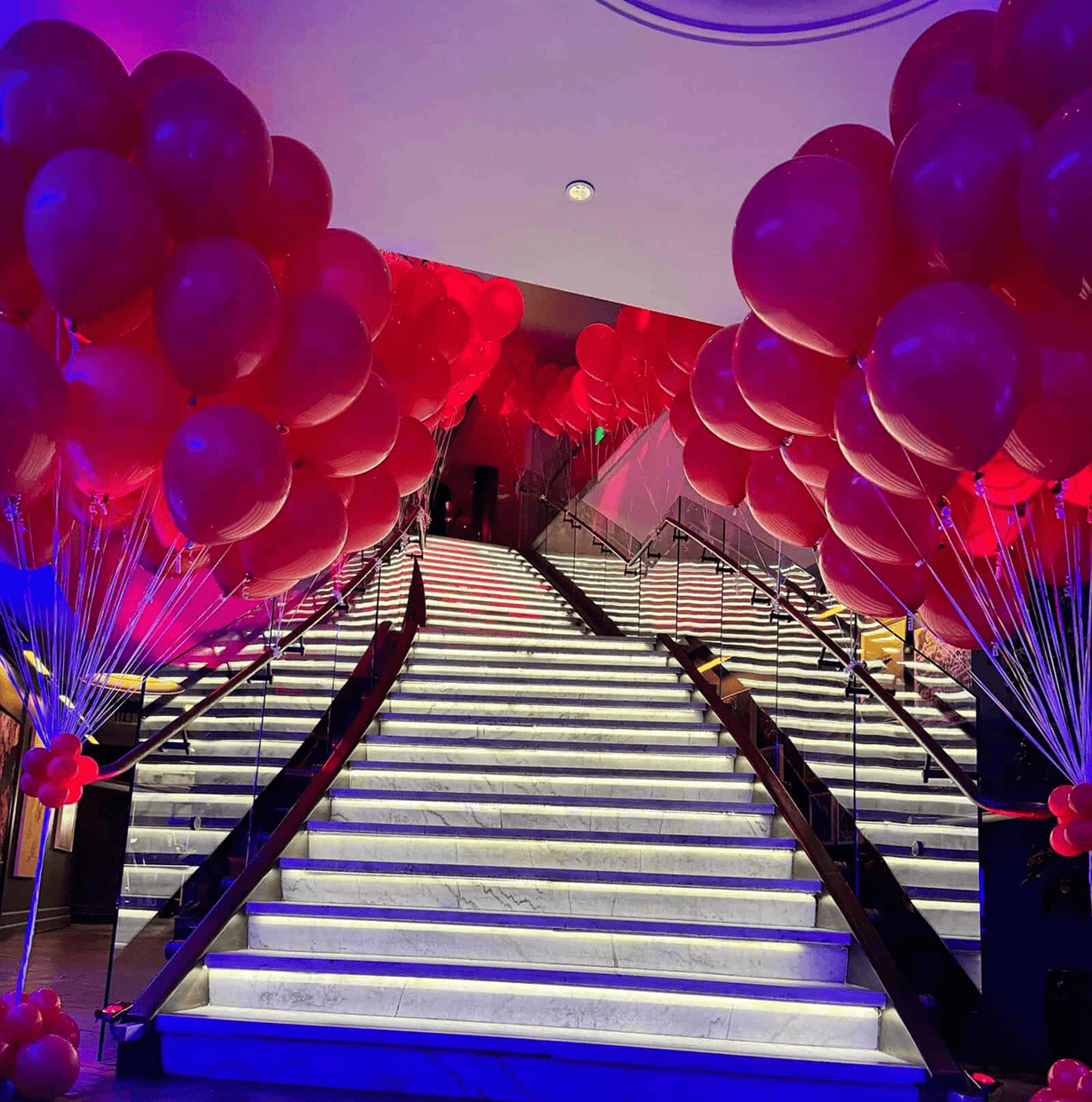 A grand marble staircase with illuminated steps is flanked by clusters of red balloons on both sides, creating a festive atmosphere.