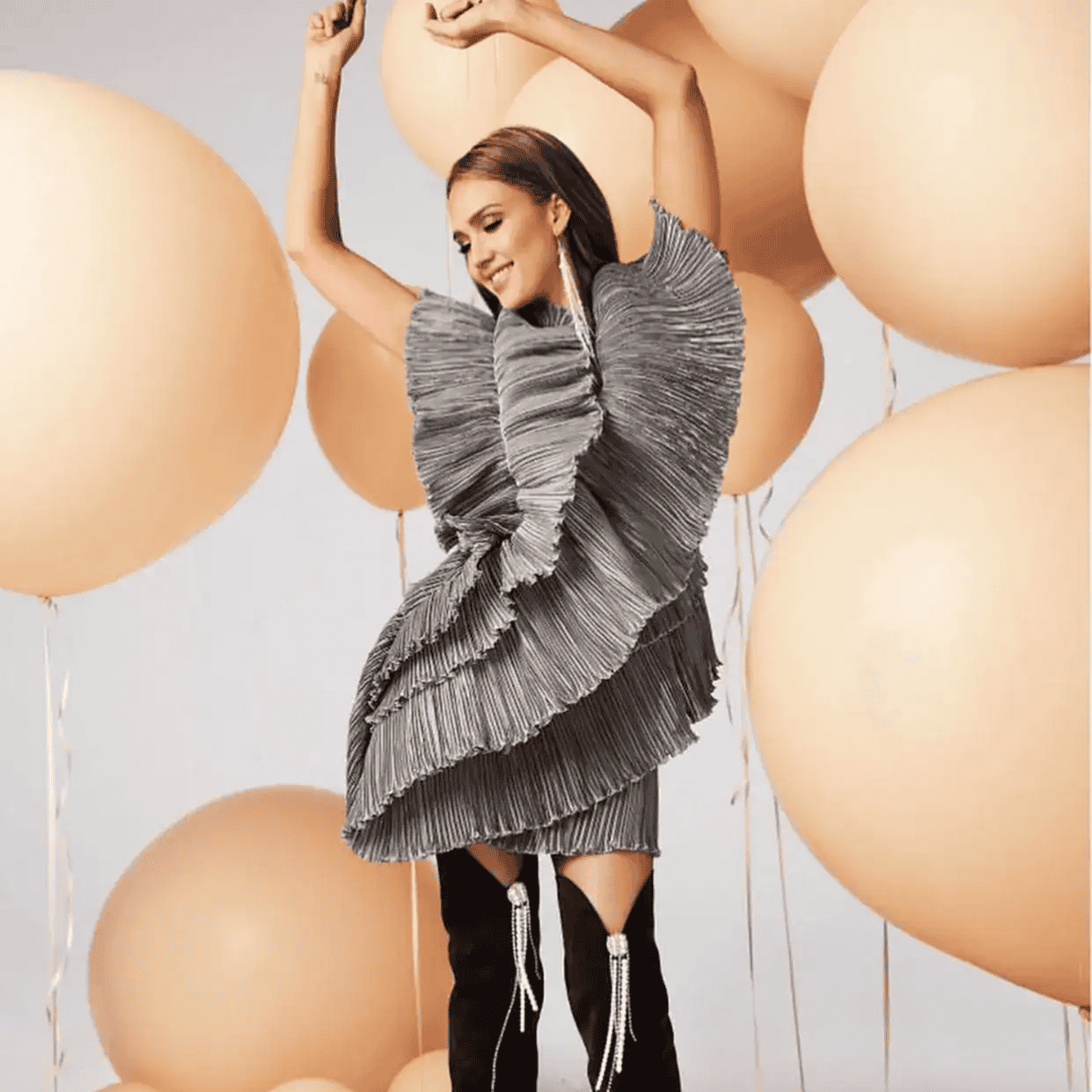 A woman poses with arms raised, wearing a ruffled, gray textured dress and black thigh-high boots, surrounded by large beige balloons.