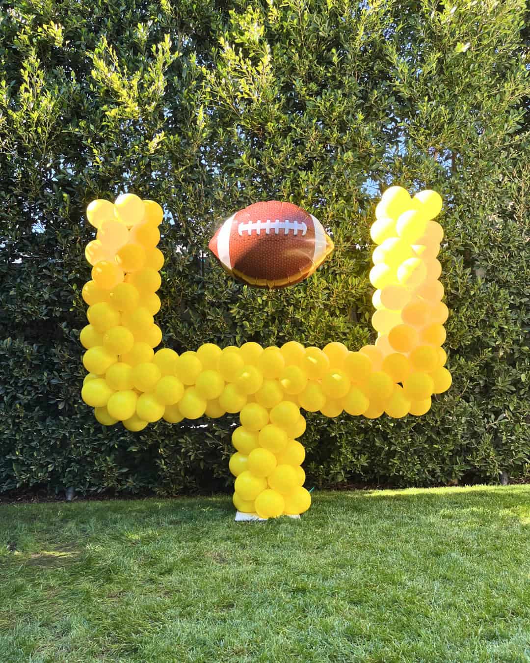 A football-shaped balloon hovers above a goalpost made of yellow balloons, creating a festive atmosphere for sporting events outdoors in front of tall green bushes.