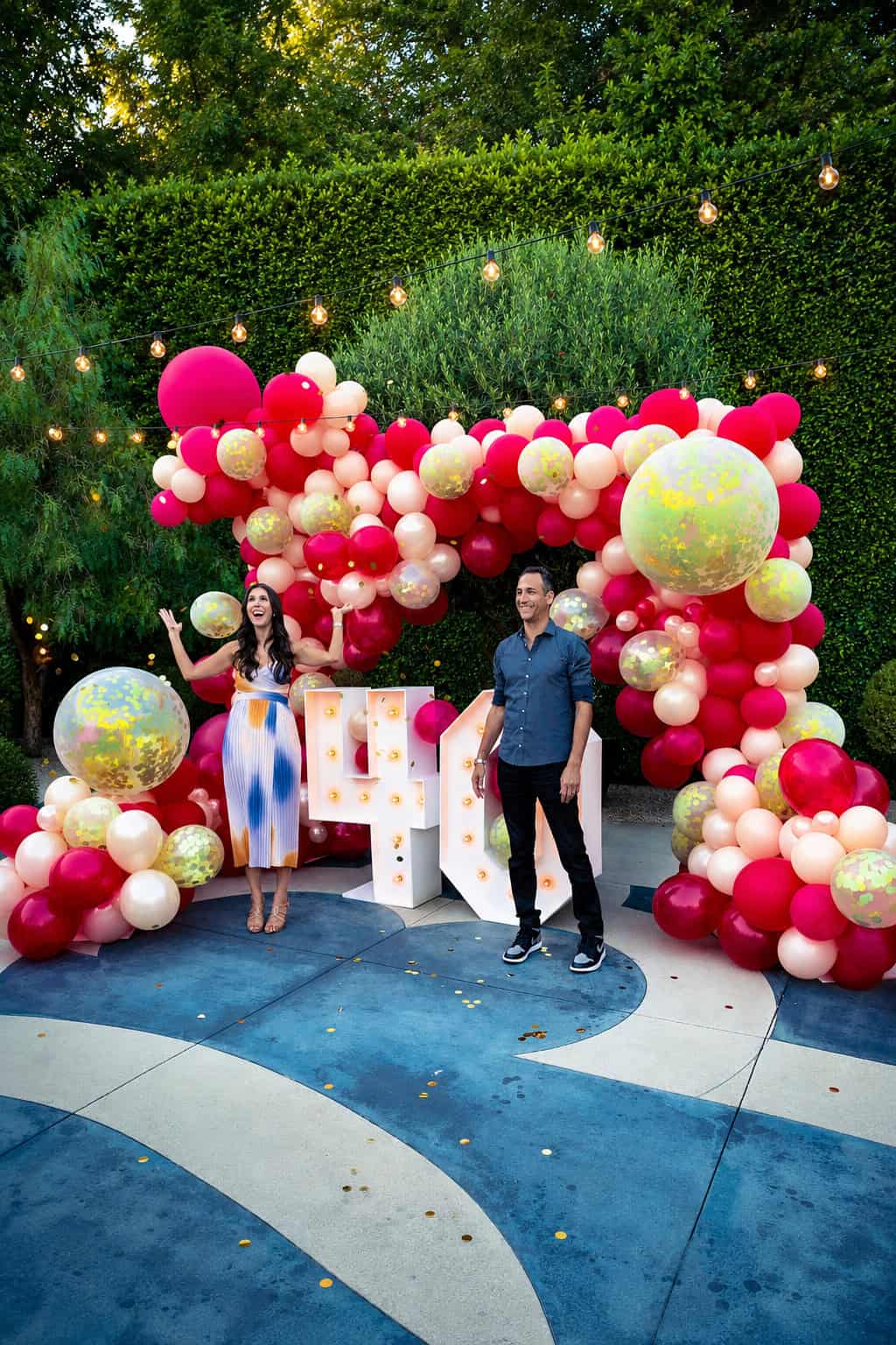 A woman and a man stand in front of a large 40 sign and a colorful pink, red, and gold balloon arch outdoors, surrounded by greenery and string lights.
