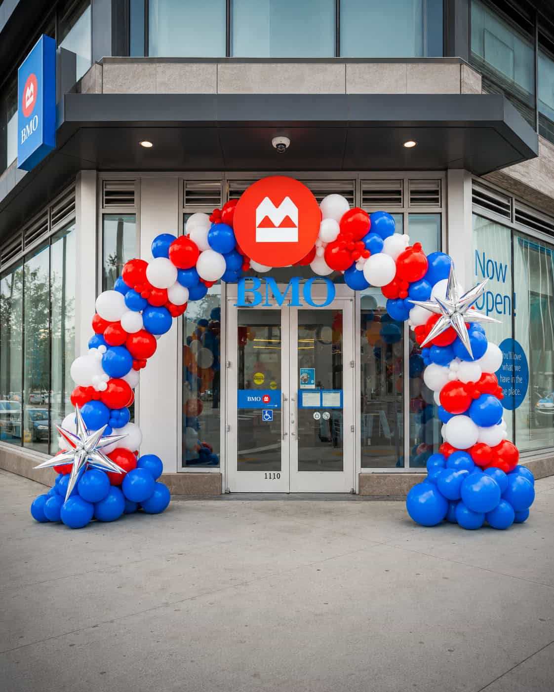 A BMO bank branch entrance decorated with a red, white, and blue balloon arch and silver star balloons, celebrating Grand Openings with a "Now Open" sign in the window.