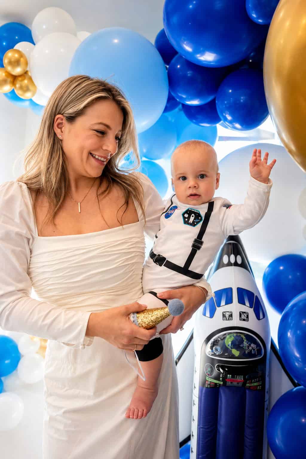 A woman in a white dress holds a baby dressed in a space-themed outfit, surrounded by blue, white, and gold balloons and a space shuttle decoration.