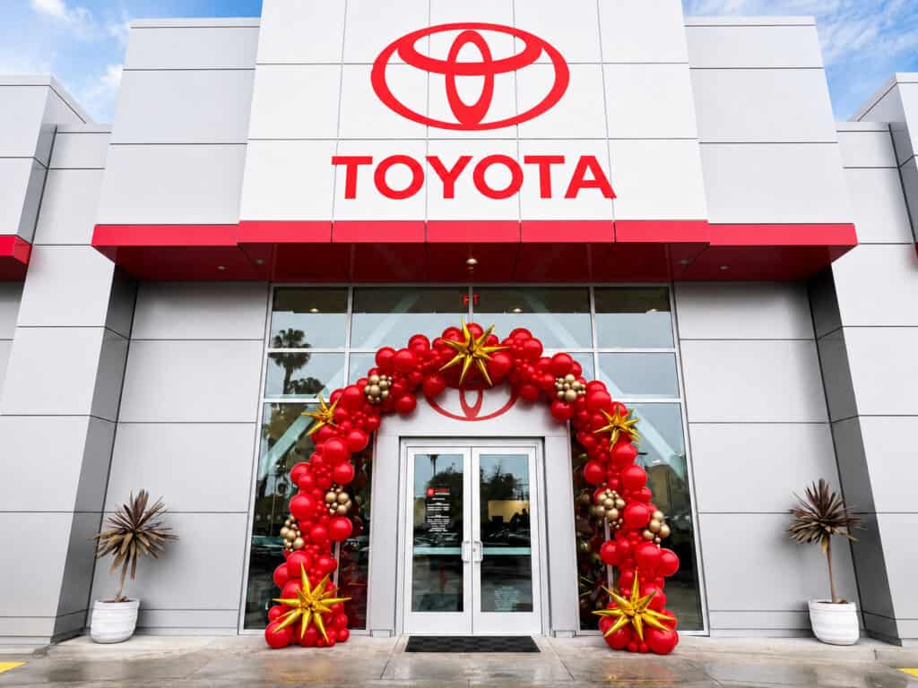 Front entrance of a Toyota dealership decorated with a red and gold balloon arch for Corporate Events, with the Toyota logo displayed above the doors.