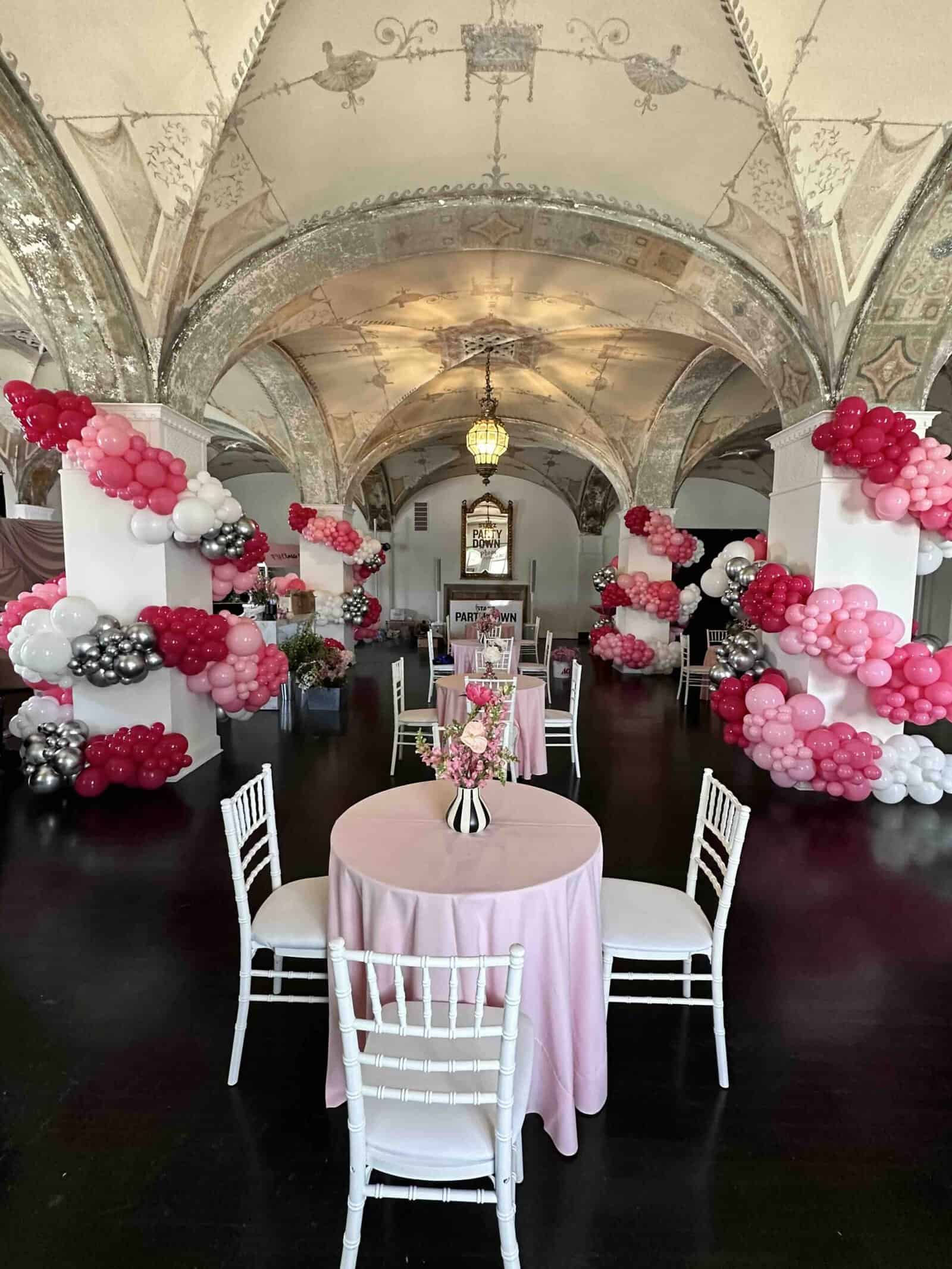Room with arched ceilings adorned with elegant balloon garlands in pink, white, red, and silver, featuring round tables with pink tablecloths and white chairs arranged for an unforgettable party decor experience.