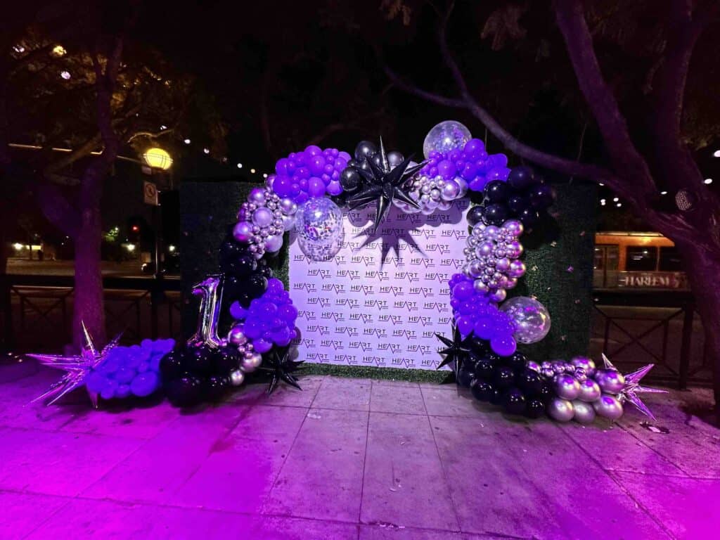 A stunning balloon arch with purple, black, and silver Balloon Decorations surrounds a step-and-repeat backdrop reading "Hey QT," featuring a silver number 1 balloon and star-shaped Event Decor accents.
