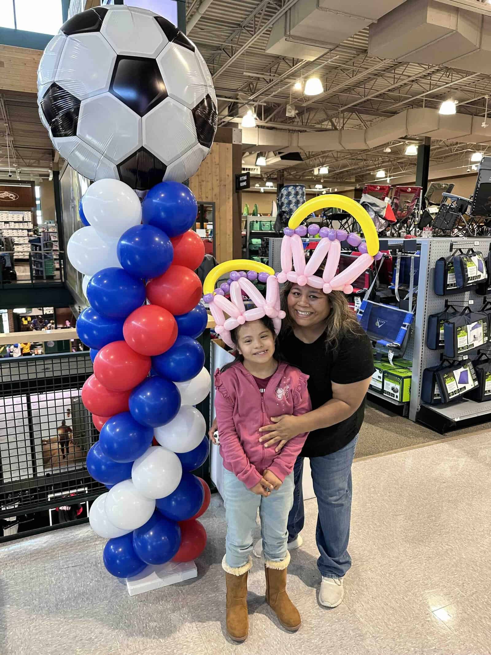An adult and a child, both wearing balloon hats made by a talented balloon artist, stand next to a tall column of red, white, and blue balloons topped with a large soccer ball inside a store.
