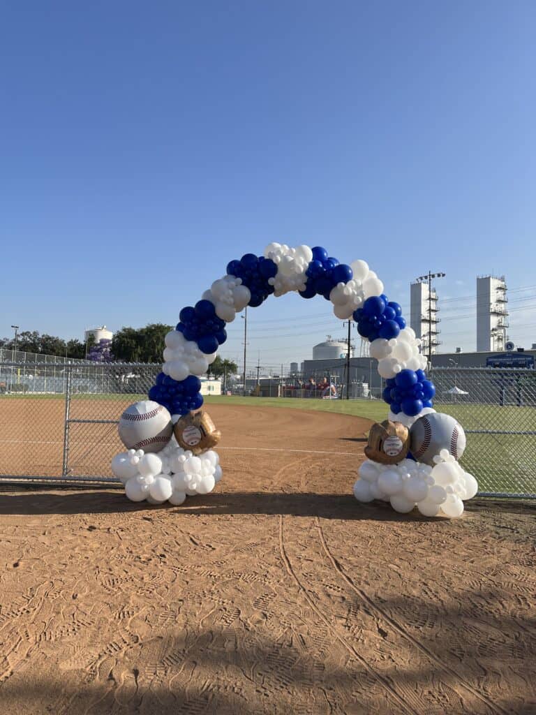 A balloon arch decorated with baseball and glove balloons stands at the entrance to a baseball field on a sunny day, showcasing festive event decor.